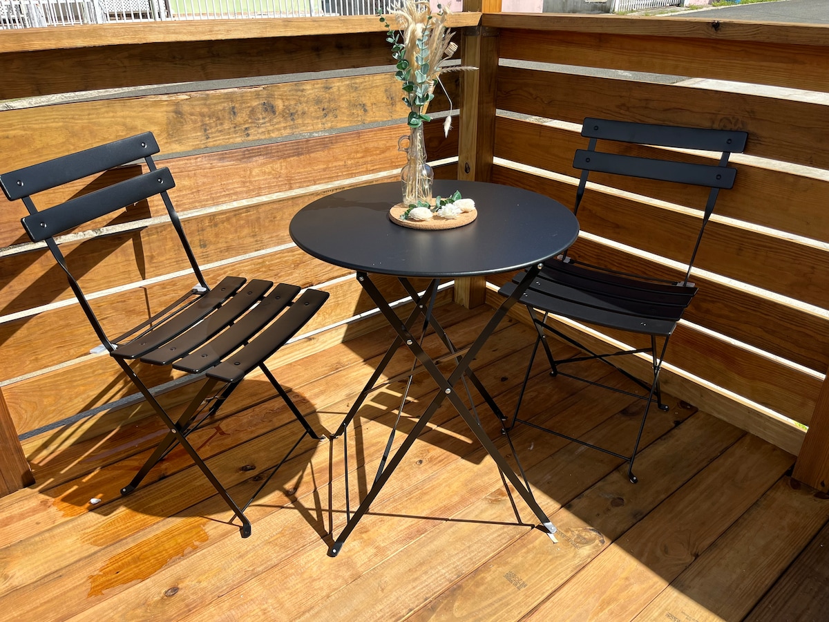 A small outdoor seating area is displayed with two black folding chairs and a round table. A simple vase holds dried flowers at the center of the table, surrounded by wooden decking that enhances the inviting space.