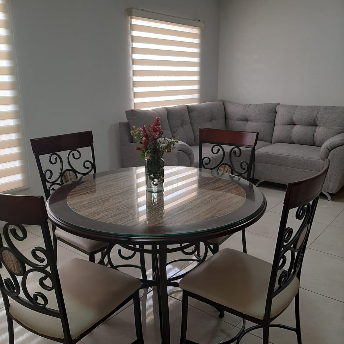 A round dining table made of glass and metal is centered in the image, surrounded by four upholstered chairs. A small vase with fresh flowers sits atop the table. In the background, a spacious gray sofa is positioned against the wall, complementing the light, neutral tones of the room.