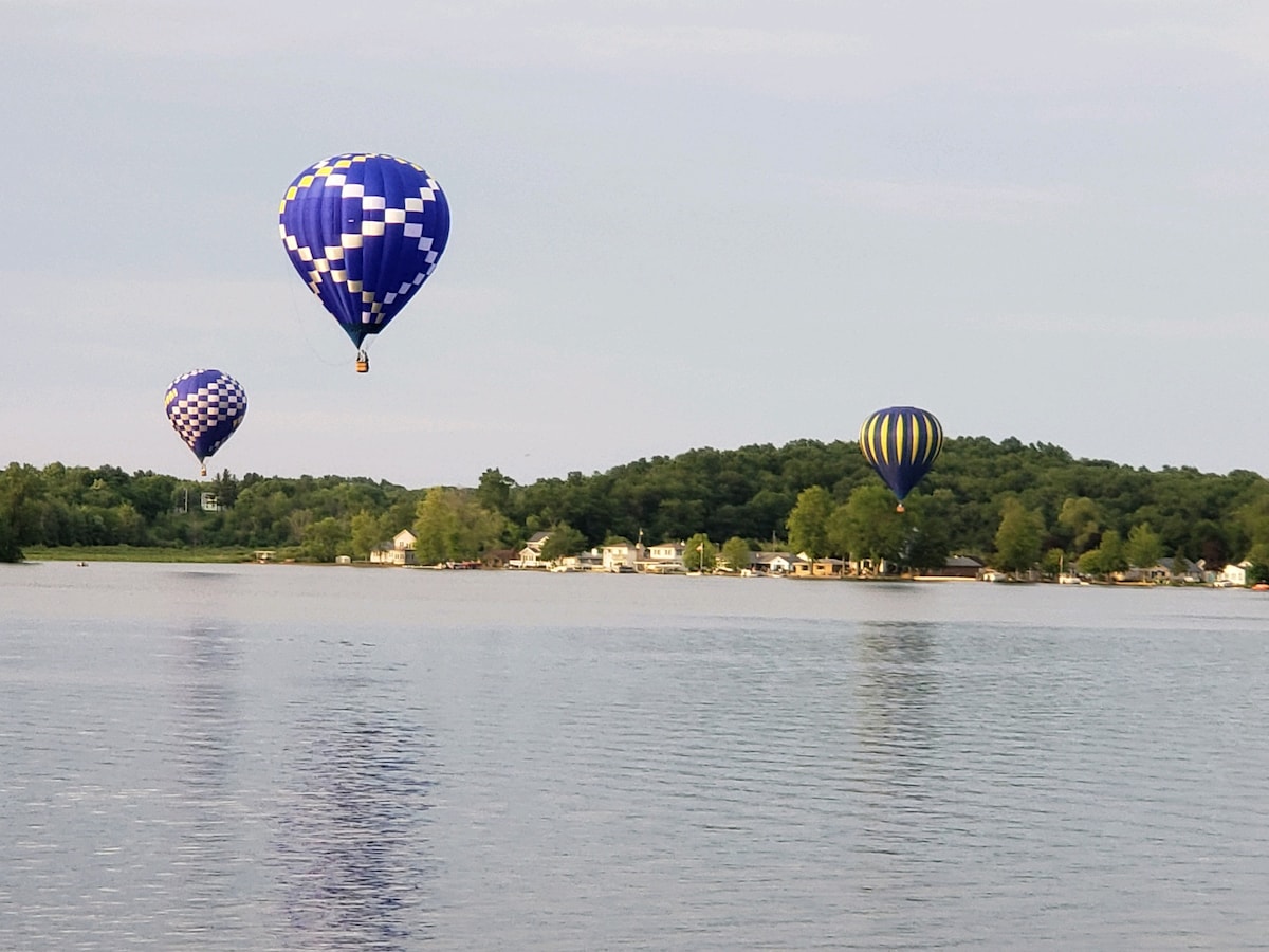 Three colorful hot air balloons float gracefully above the calm waters of the lake, contrasting with the lush greenery of the shoreline. A small cluster of houses is visible in the background, nestled among the trees, reflecting the peaceful atmosphere of the scene.