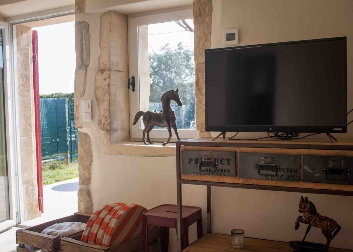 A rustic display is presented with a mounted television above a wooden console table. A decorative horse figurine stands on the windowsill, while sunlight illuminates the room through a nearby window, enhancing the texture of the stone walls.