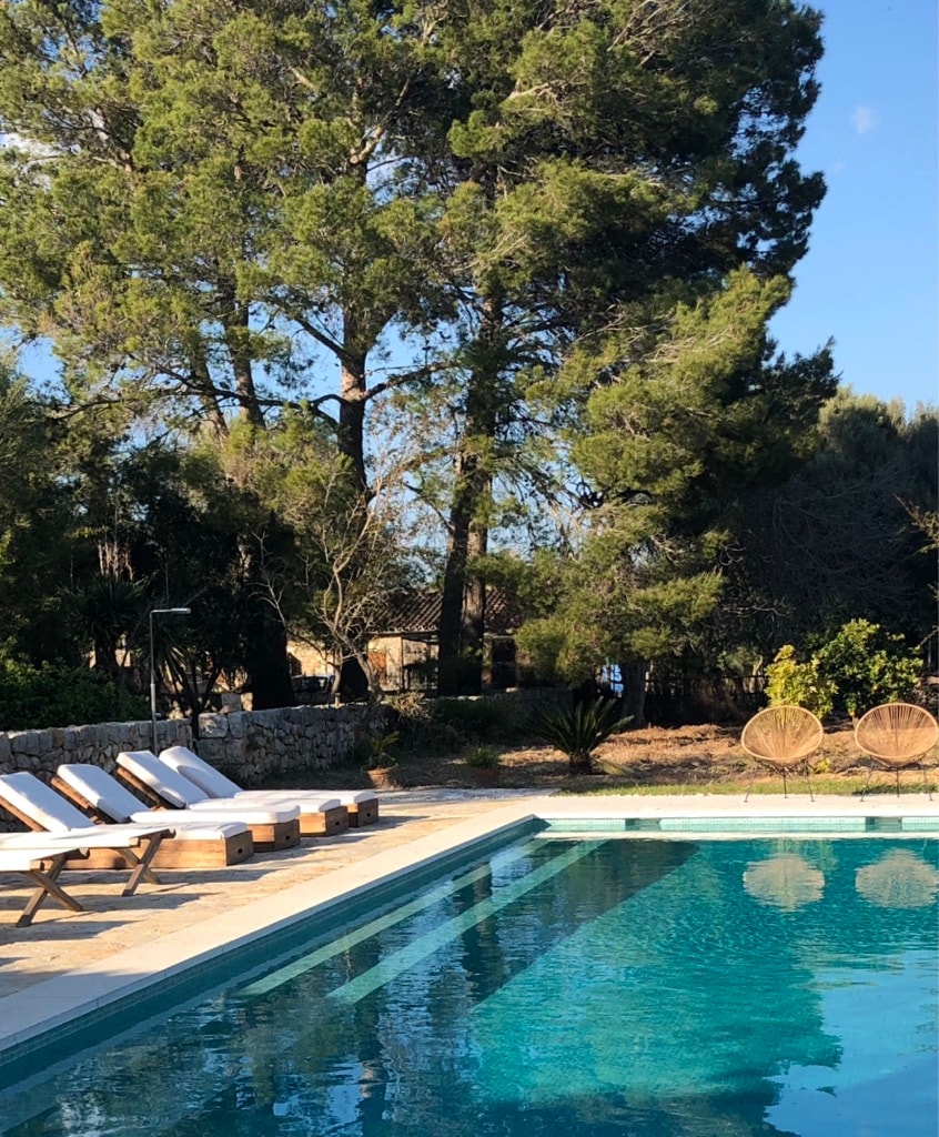 A serene swimming pool is framed by tall trees and lush greenery, offering a refreshing view. Sun loungers are positioned along the poolside, inviting relaxation. The clear water reflects the sunlight, enhancing the outdoor experience.