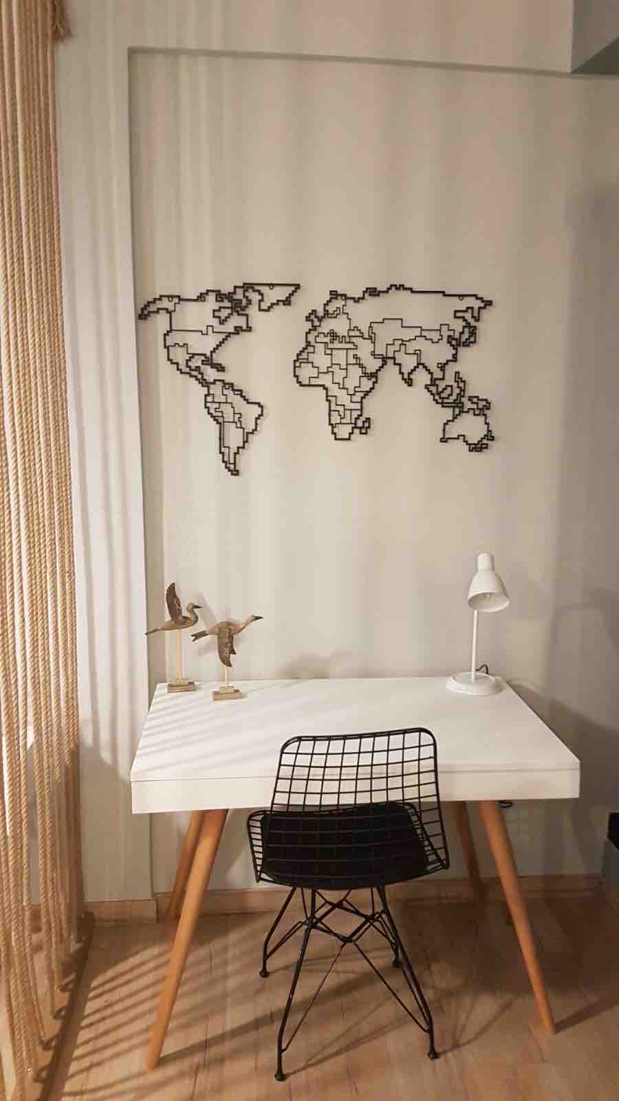 A minimalist workspace is presented featuring a white desk and a wire chair. A small desk lamp illuminates the area. A decorative wire map of the world hangs on the wall, adding an artistic touch to the space.