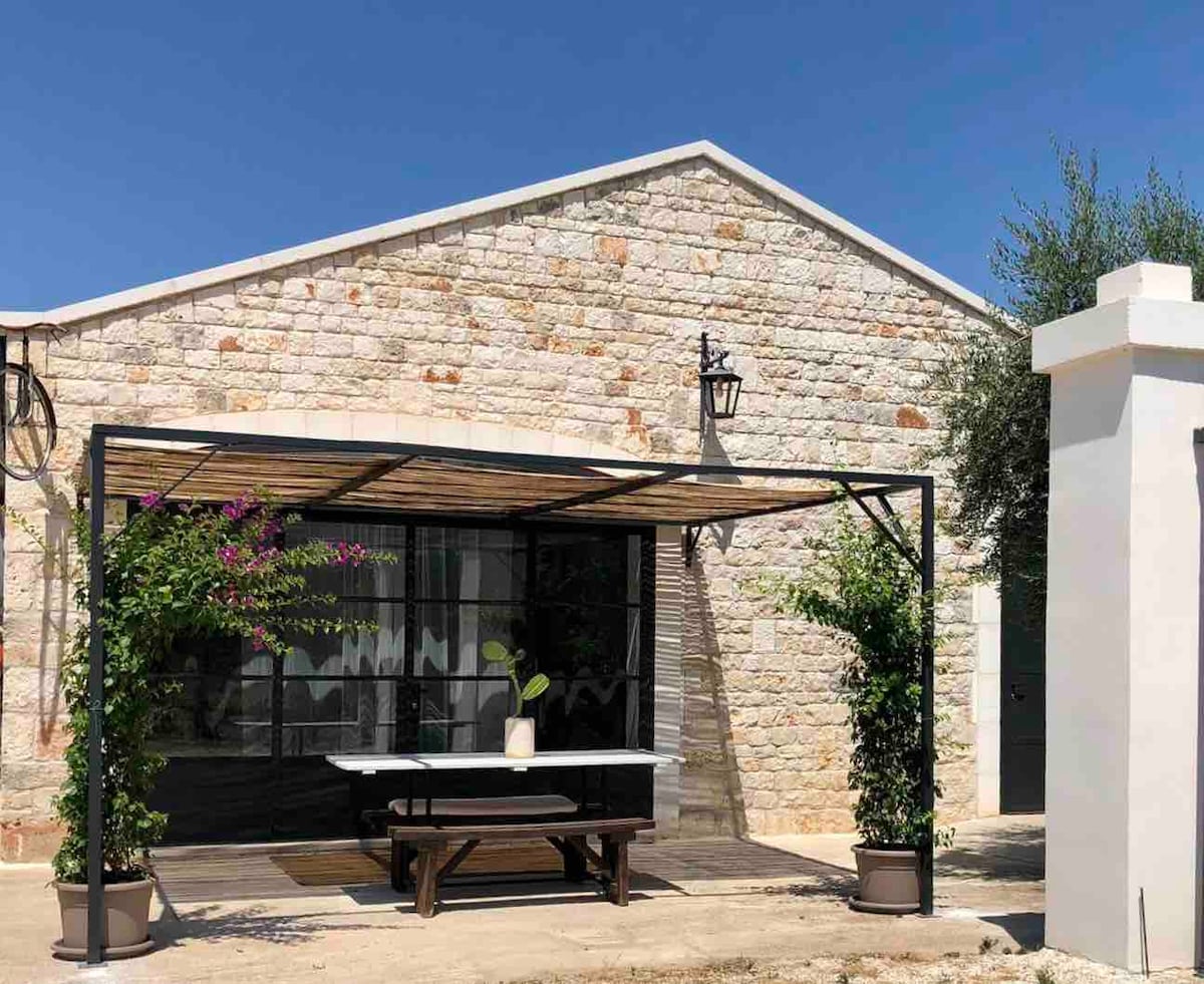 An outdoor seating area features a large table beneath a pergola, surrounded by climbing plants. A stone structure serves as the backdrop, with large glass doors allowing visibility into the interior. Natural light enhances the inviting atmosphere of the space.