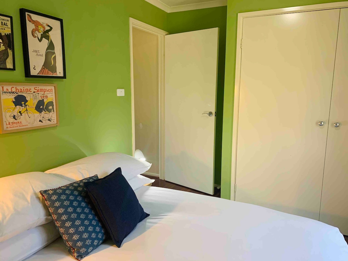 A compact sleeping area features a spacious bed with neatly arranged white linens and decorative pillows. Bright green walls provide a lively backdrop, while framed artwork adorns the space. A light-colored door leads to another room, complementing the room's overall bright and welcoming appearance.