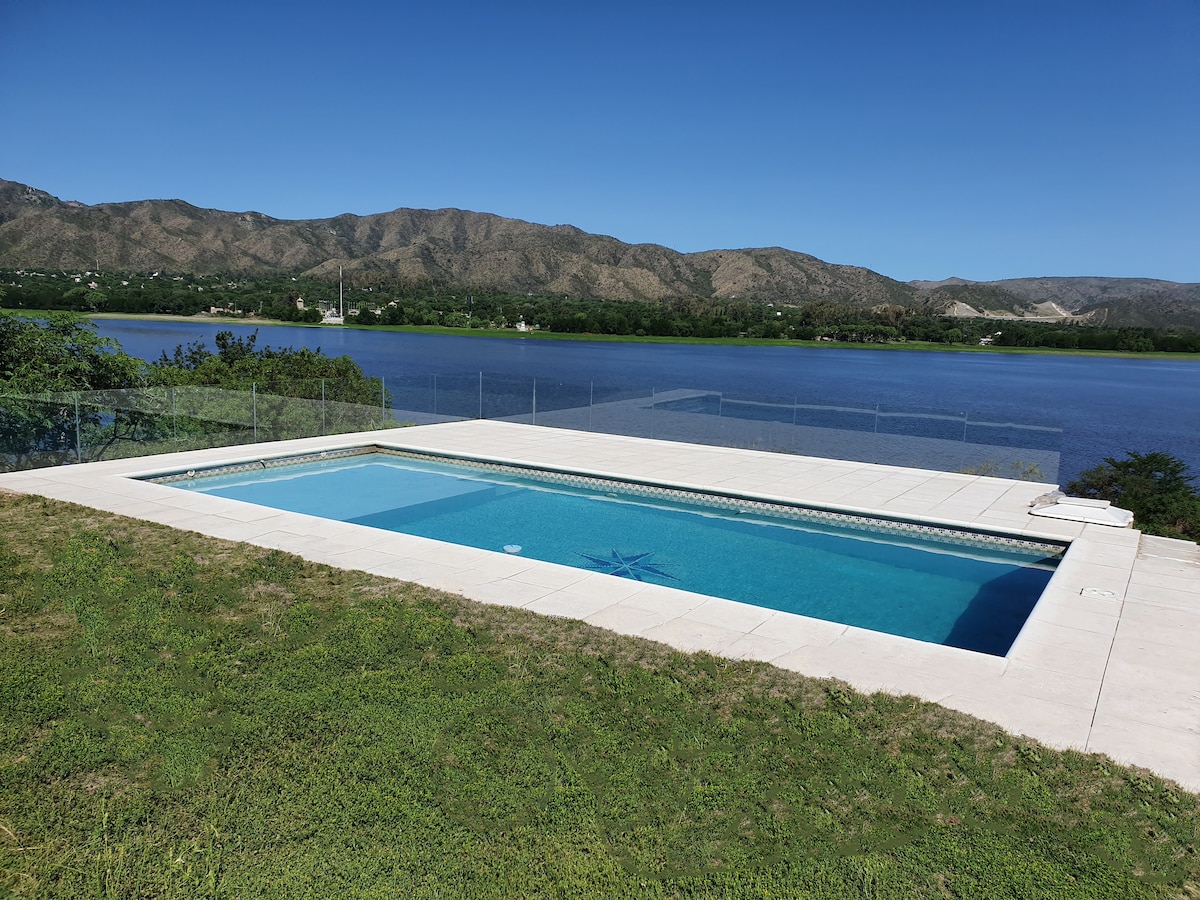 An outdoor swimming pool is showcased, surrounded by a paved patio and lush grass. The pool offers unobstructed views of the lake and distant hills, providing a serene backdrop, with clear blue skies overhead.
