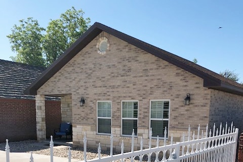 A charming exterior view of the Casita features a light brick facade complemented by large windows. The entrance is framed by a white decorative fence, with a small seating area visible on the porch, inviting a sense of comfort and openness.