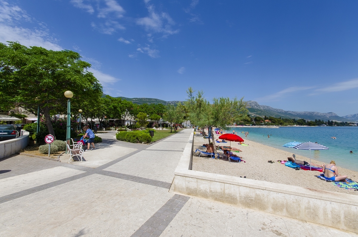 A vibrant beachfront promenade is lined with trees and benches, leading to a sandy beach. Colorful umbrellas dot the shore while individuals relax under the sun beside the calm, clear water. The distant mountains provide a picturesque backdrop to the seaside scene.