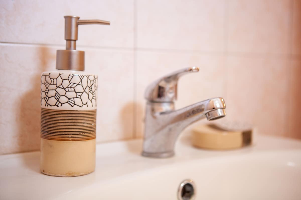 A stylish soap dispenser is positioned on the edge of a sink, displaying a modern design with a geometric pattern. A shiny chrome faucet complements the dispenser, reflecting light. The background features neutral-toned tiled walls, contributing to a clean and functional bathroom ambiance.