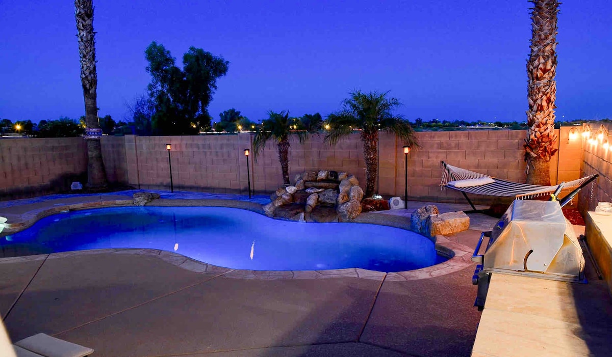 An inviting pool area features a clear, blue pool complemented by rock formations and tropical plants. Two palm trees provide shade, and a comfortable hammock is nearby. The area is softly illuminated by lights, creating a serene atmosphere suitable for relaxation in the evening.