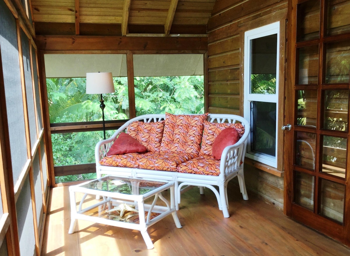A screened-in porch features a white wicker sofa adorned with tropical-patterned cushions and red accent pillows. A small glass coffee table sits in front, and a floor lamp provides soft illumination. Natural light filters through the surrounding screens, highlighting the lush greenery outside.