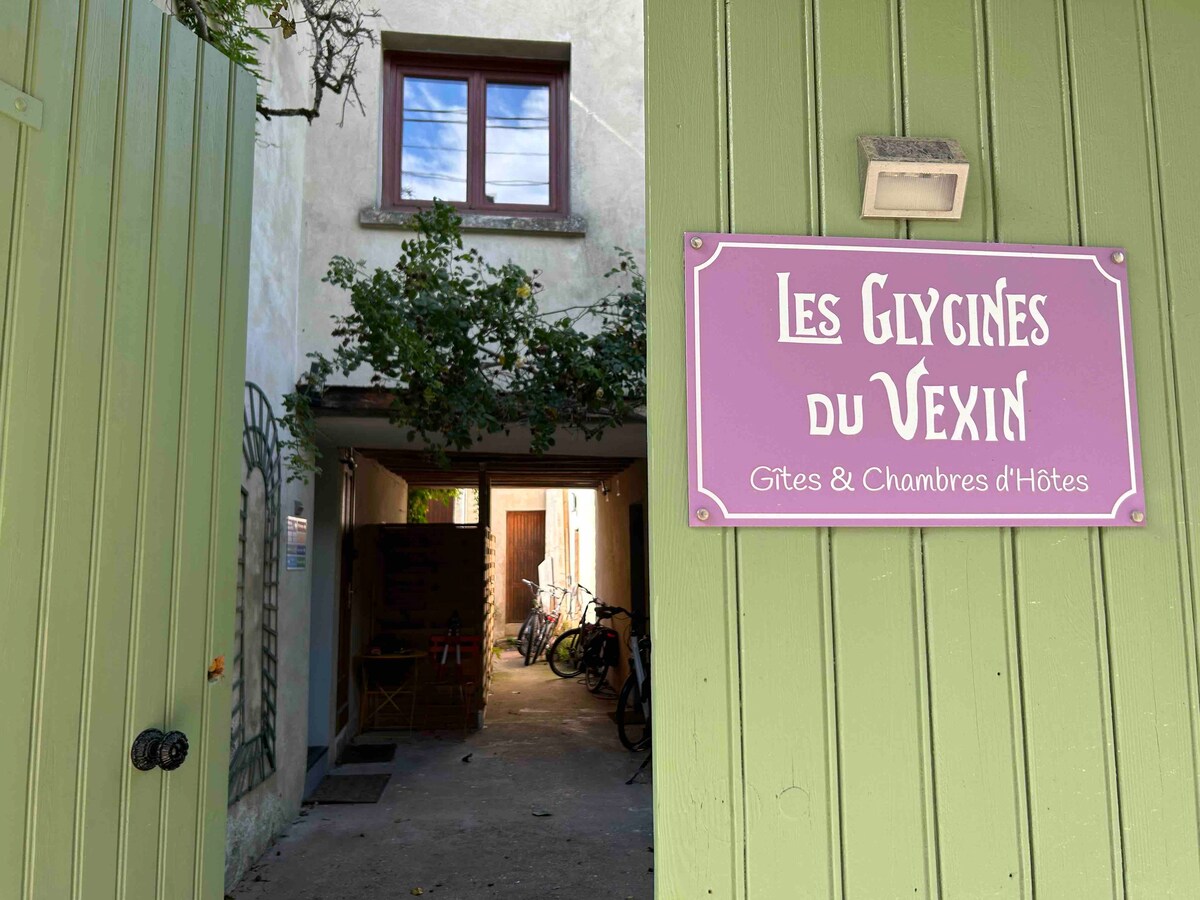 A green wooden gate leads to an inviting pathway, framed by the rustic charm of the building. A sign reading 'Les Glycines du Vexin' is prominently displayed. Bicycles are parked along the side, hinting at a welcoming atmosphere for guests.