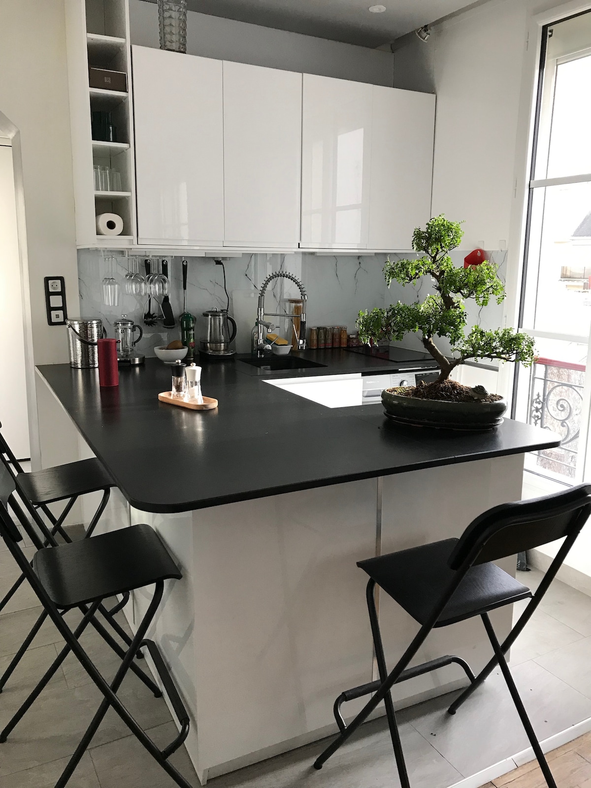 An open kitchen area features a black countertop with high stools arranged around it. Modern cabinets above and below provide storage space, while various kitchen appliances are visible. A decorative bonsai tree sits on the countertop, adding a touch of greenery to the space.