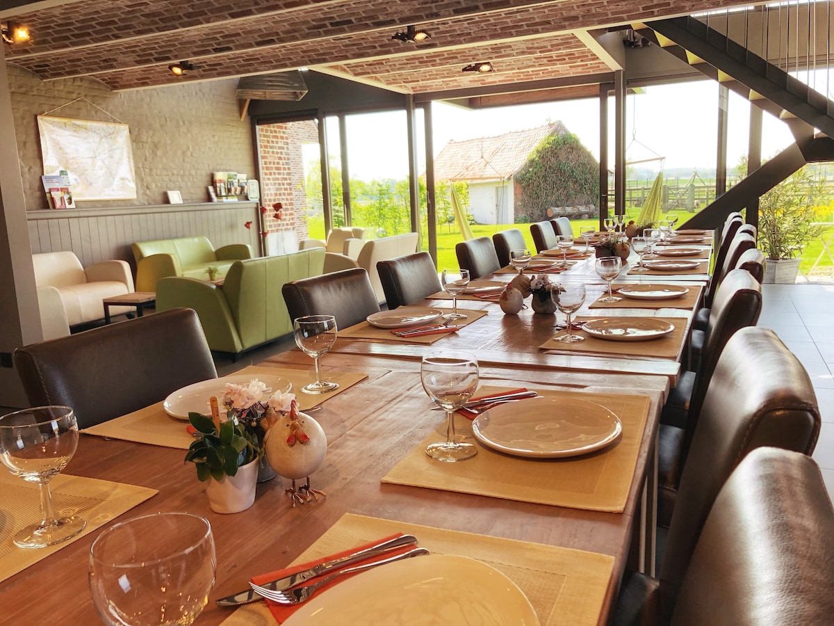 A spacious dining area features a large wooden table set with plates and glassware. Natural light streams through expansive windows, providing picturesque views of the garden and fields. Comfortable seating is arranged nearby, enhancing the inviting atmosphere.