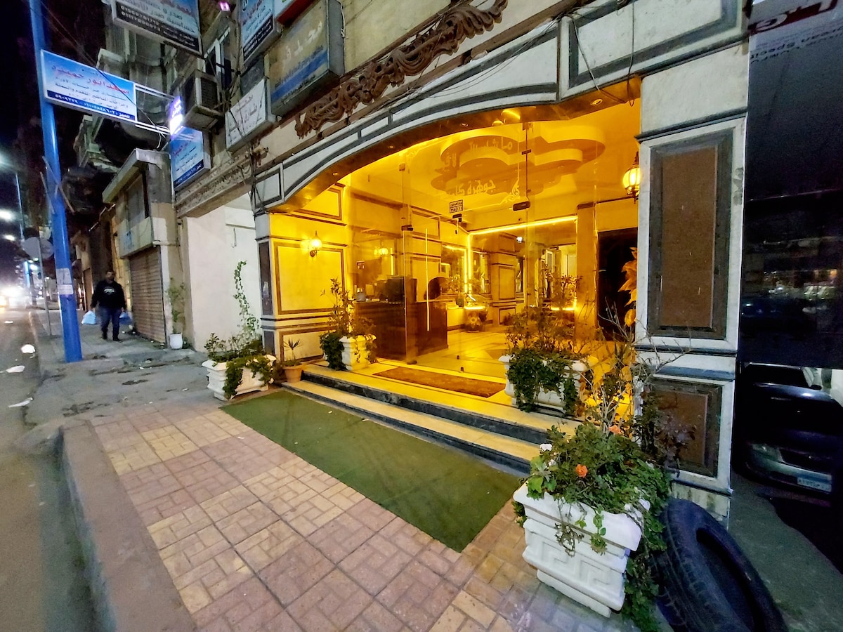The entrance of the building features a large glass façade, illuminated with warm lighting. Potted plants line the walkway, enhancing the welcoming atmosphere. The surrounding area includes a paved pathway and street signage, reflecting the bustling neighborhood.