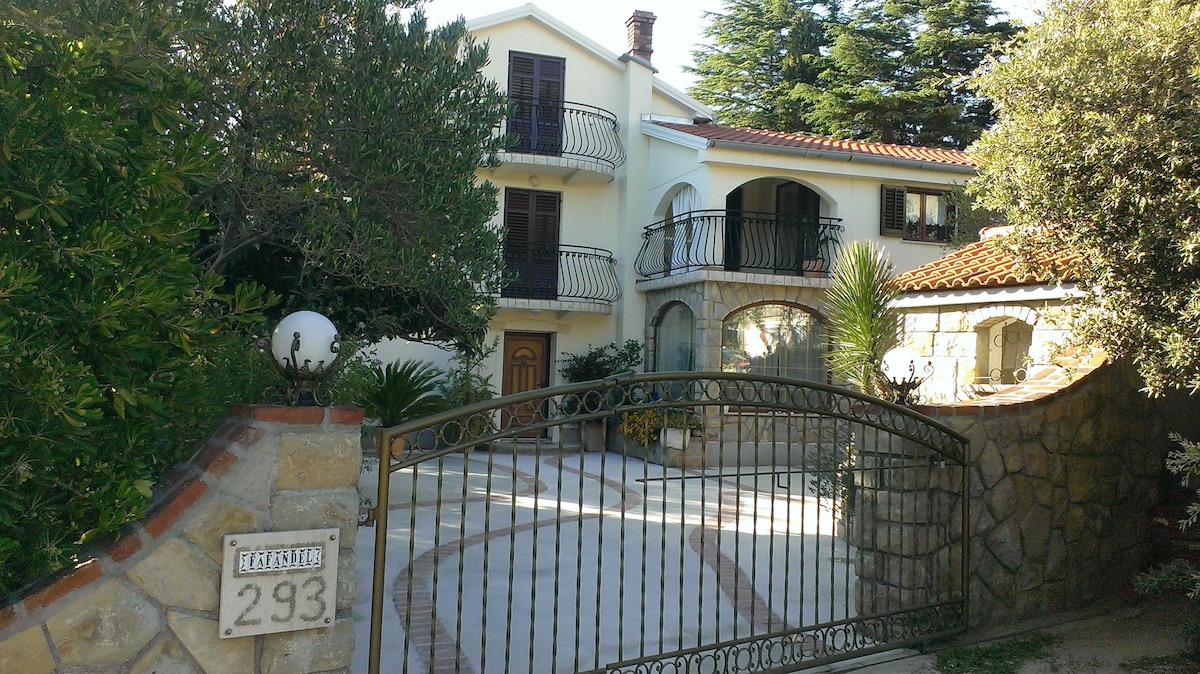 The exterior of the property is shown, featuring a decorative stone entrance gate and a spacious driveway. The house displays an elegant architectural style with balconies on the upper level and lush greenery surrounding the structure.