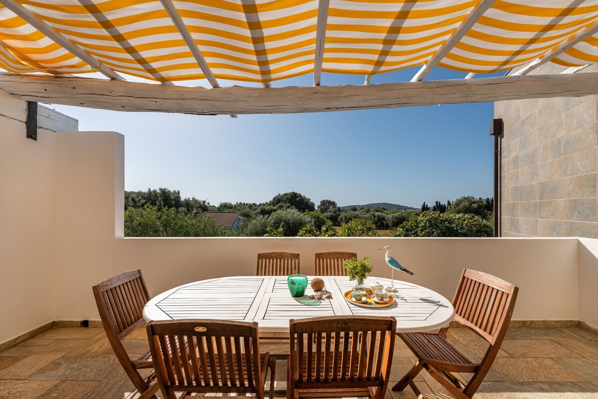 A spacious outdoor dining area is set under a striped canopy, featuring a round table surrounded by wooden chairs. A view of lush greenery is visible in the background, with the horizon gently fading into the distance.