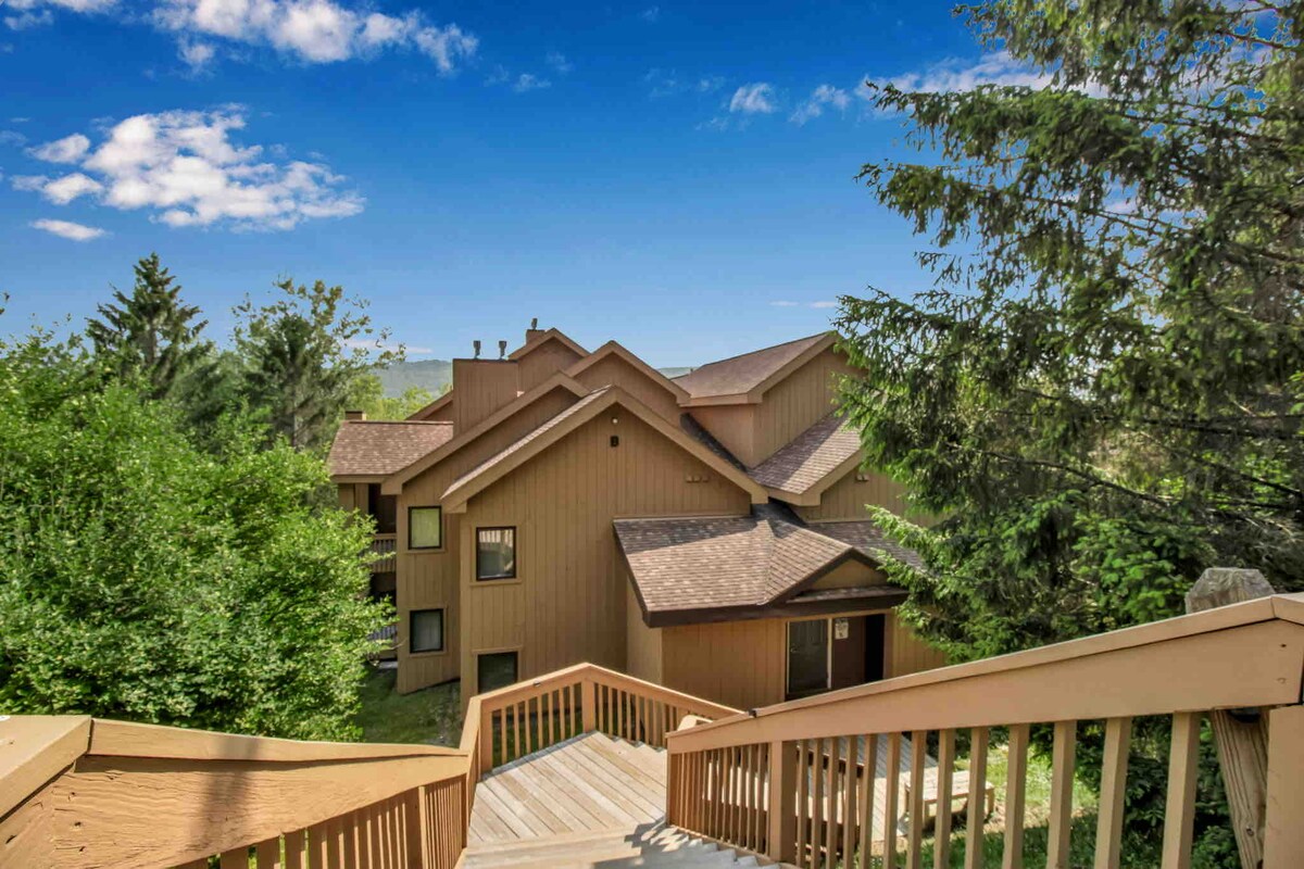 A wooden staircase leads down through a lush green landscape towards a multi-unit building. The building features a mix of large windows and wood siding, surrounded by trees and under a clear blue sky.