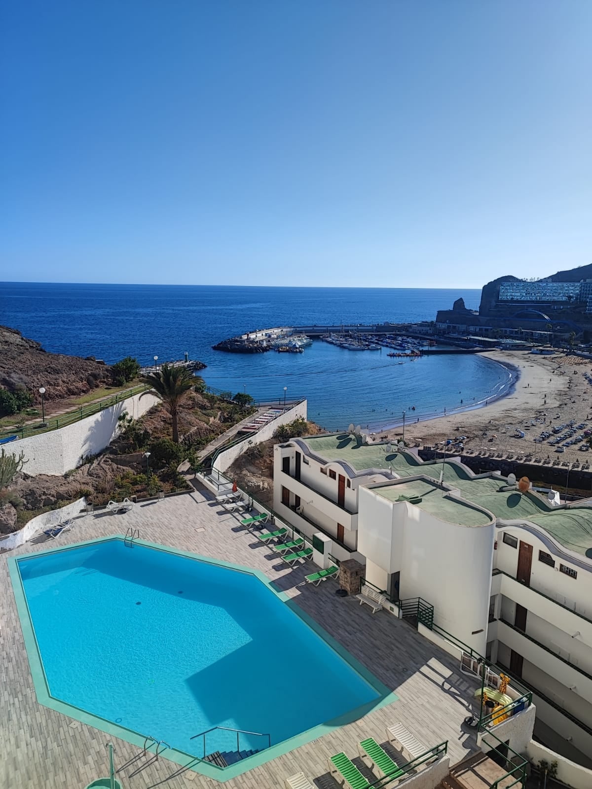 An inviting community pool is situated at the forefront, overlooking a sandy beach and a calm blue sea. The area is framed by lush palm trees and gentle hills, while structures with a clean white facade accompany the scenic view.
