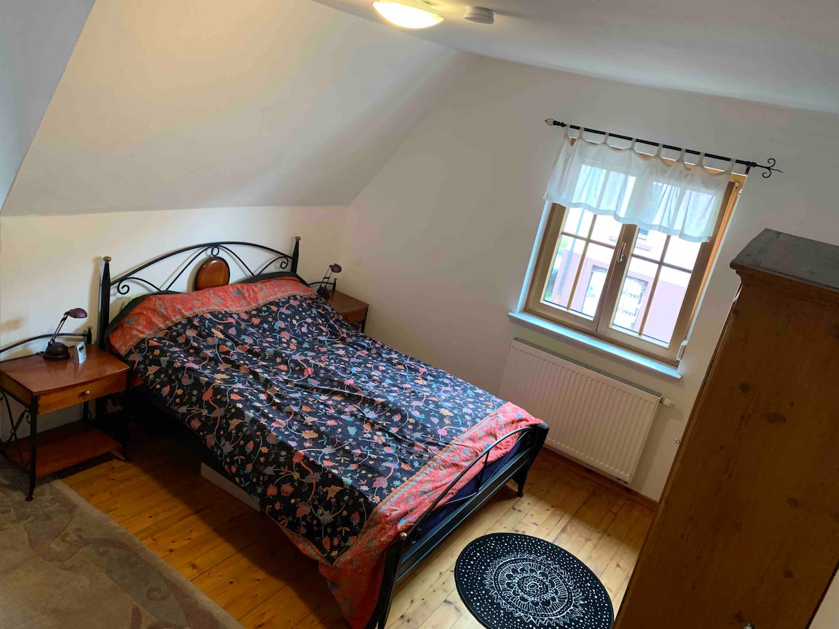 A cozy sleeping area features a spacious bed covered with a patterned blanket. Two bedside tables with lamps flank the bed. A window with sheer curtains allows natural light to enter, illuminating the wooden floor and the decorative rug beneath.