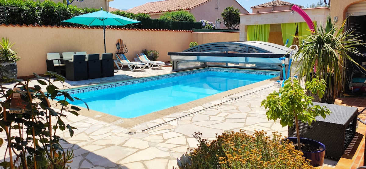 A serene swimming pool, covered and heated, is framed by a spacious stone patio. Sun loungers are positioned next to the pool, with a shaded seating area under a vibrant umbrella. Lush greenery and colorful flowers enhance the inviting outdoor setting.