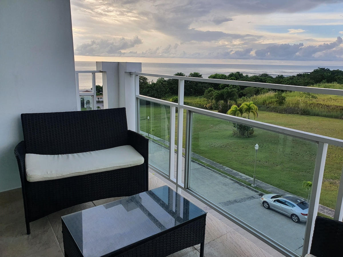 A private balcony area is visible, featuring a dark wicker seating arrangement with a light cushion and a glass-top table beneath. The view extends toward open green spaces and the ocean, with soft clouds reflecting on the water's surface, enhancing a sense of tranquility.