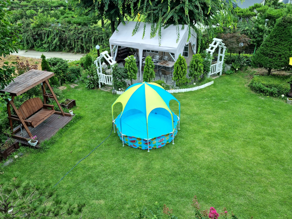 A spacious lawn garden is featured, highlighted by a colorful tent in the center. Surrounding the area are neatly trimmed hedges and decorative features, including a wooden swing and a gazebo, providing a serene outdoor space for relaxation.