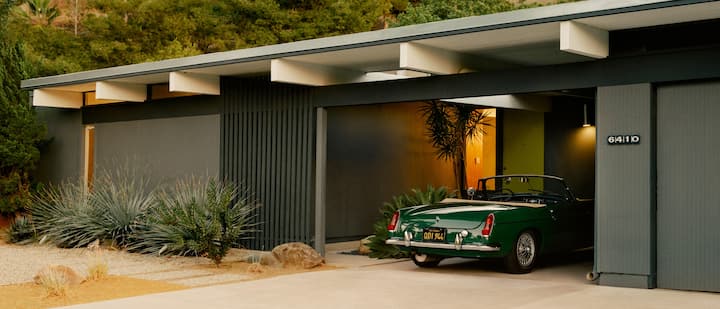 Une photo montre l'extérieur d'une maison de plain-pied dans le désert, dotée d'une place de parking couverte où se trouve une voiture de sport classique décapotable.