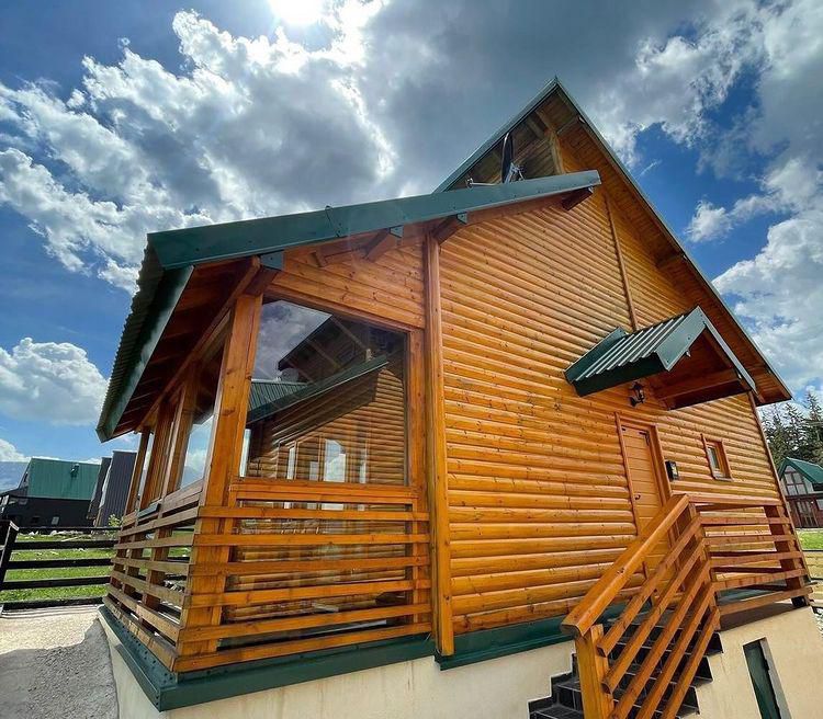 A wooden cabin is shown with a sloped roof and large windows, allowing natural light to fill the interior. A spacious porch with wooden railings is visible, providing an inviting entryway. The sky features scattered clouds, contributing to a serene atmosphere.