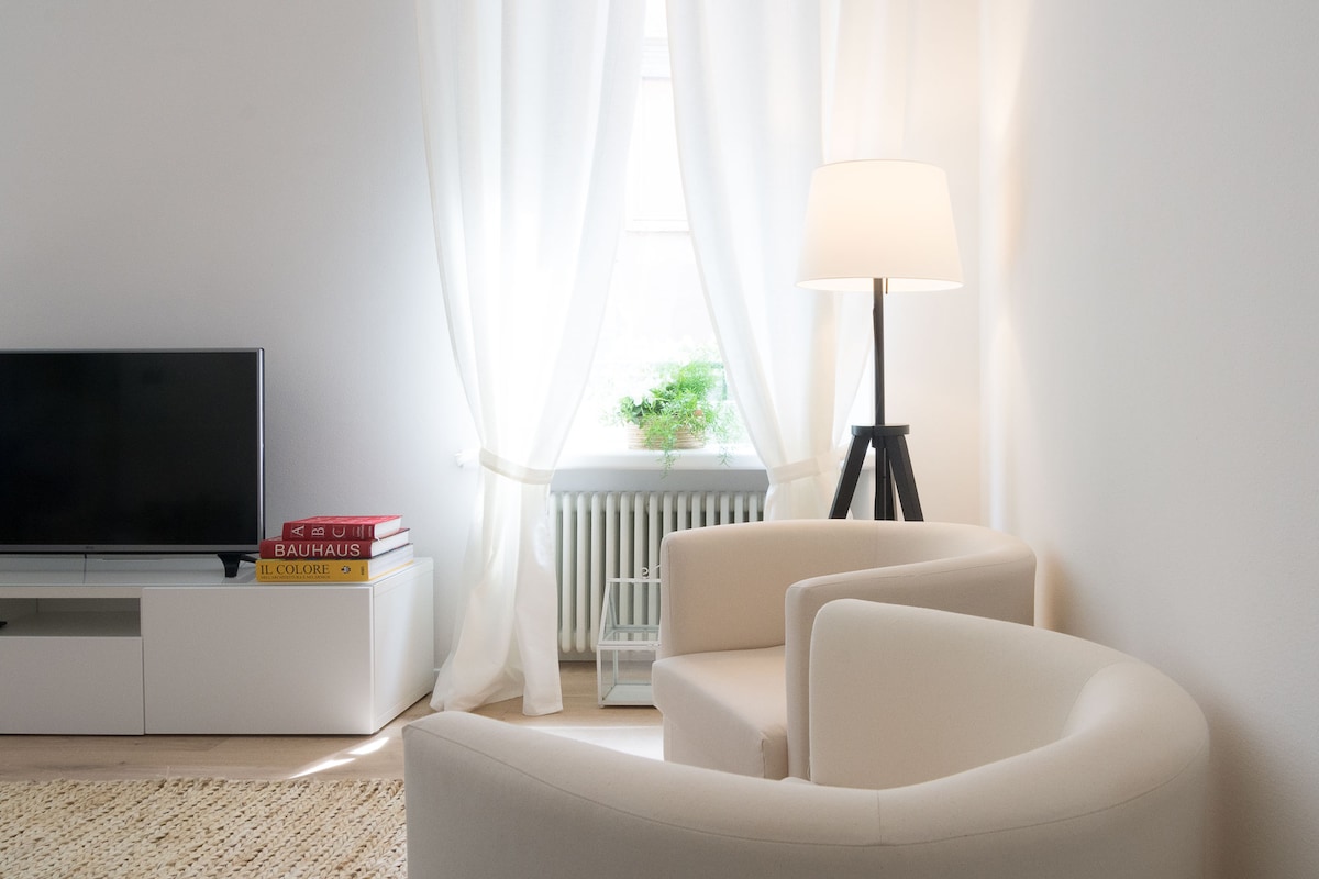 A bright living area is depicted with soft white walls and natural light filtering through sheer curtains. Two modern, curved armchairs are positioned beside a simple lamp. A flat-screen television and minimalist media console create a clean and inviting space.