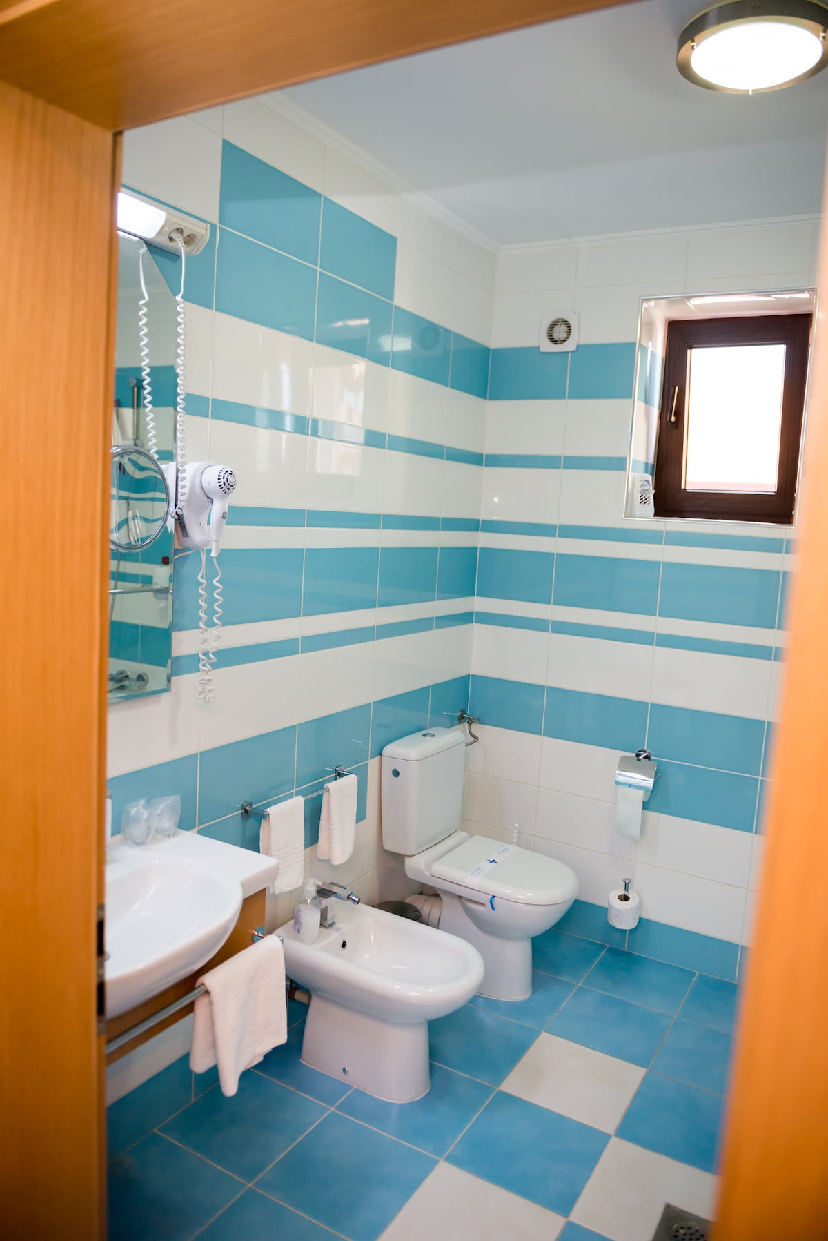 The bathroom features a bright color scheme with blue and white tiles. A wall-mounted hairdryer is visibly positioned above the sink, and essential toiletries are neatly arranged. Two flush toilets and a bidet are present, along with neatly hung towels by the sink.