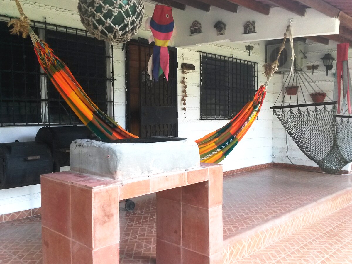 Two vibrant hammocks are suspended on a covered terrace, providing a relaxed seating area. A concrete barbecue grill is positioned nearby, with decorative wall hangings and potted plants visible. The space is framed by large windows with protective bars, enhancing a cozy and inviting environment.