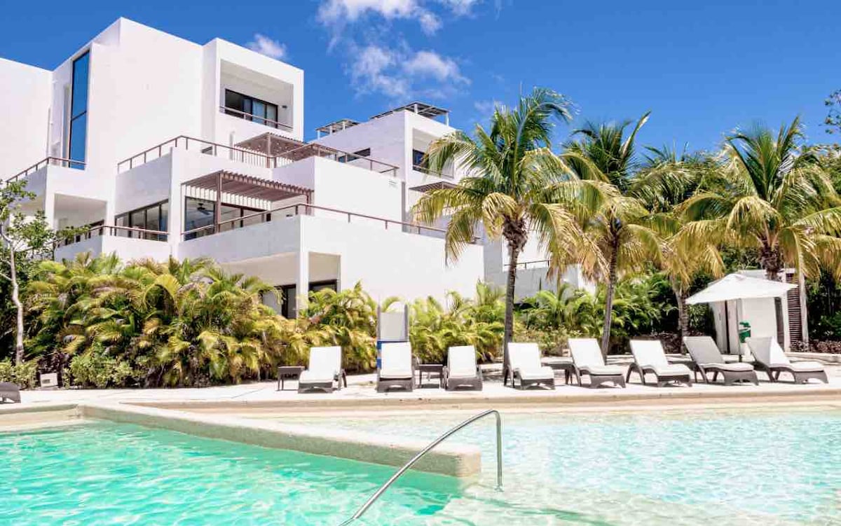 A modern apartment building is surrounded by lush greenery and palm trees. Lounge chairs are positioned by a crystal-clear pool, inviting guests to relax. A shaded cabana provides additional seating for outdoor enjoyment under the bright blue sky.