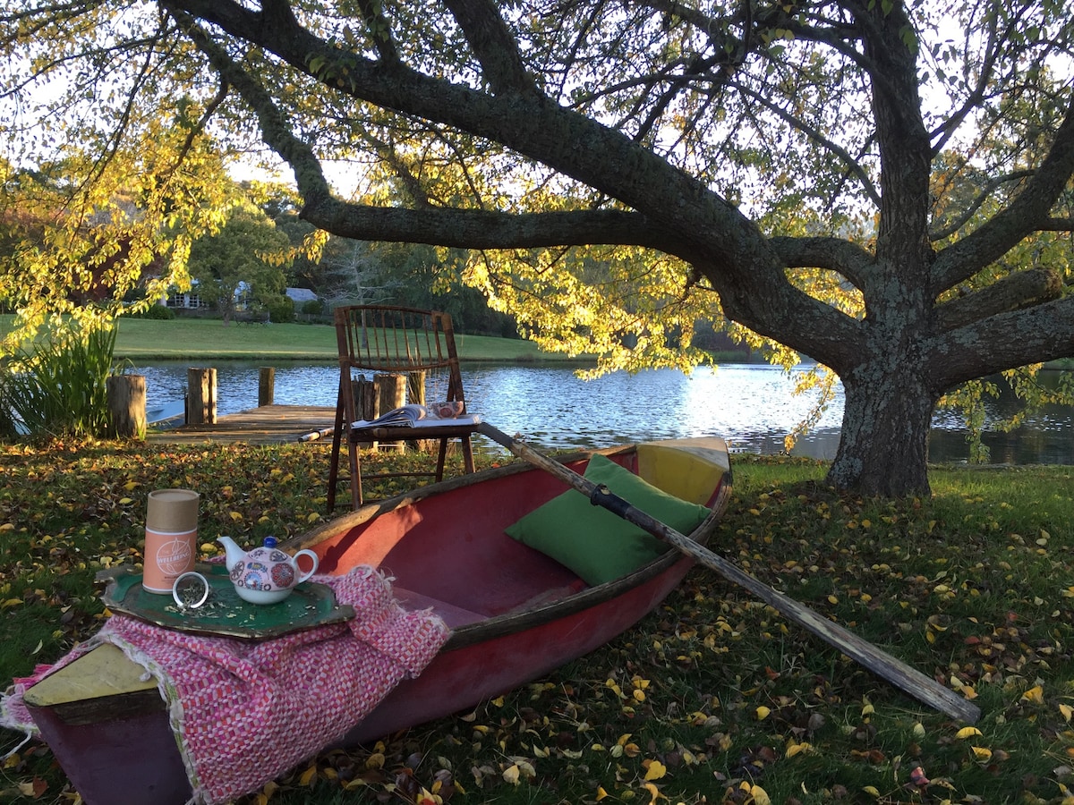 A rustic rowing boat rests on a grassy bank beneath a large tree, with golden leaves scattered around. A comfortable chair is positioned nearby, along with a small table set with a tea service and cushions, creating a peaceful setting by the water.