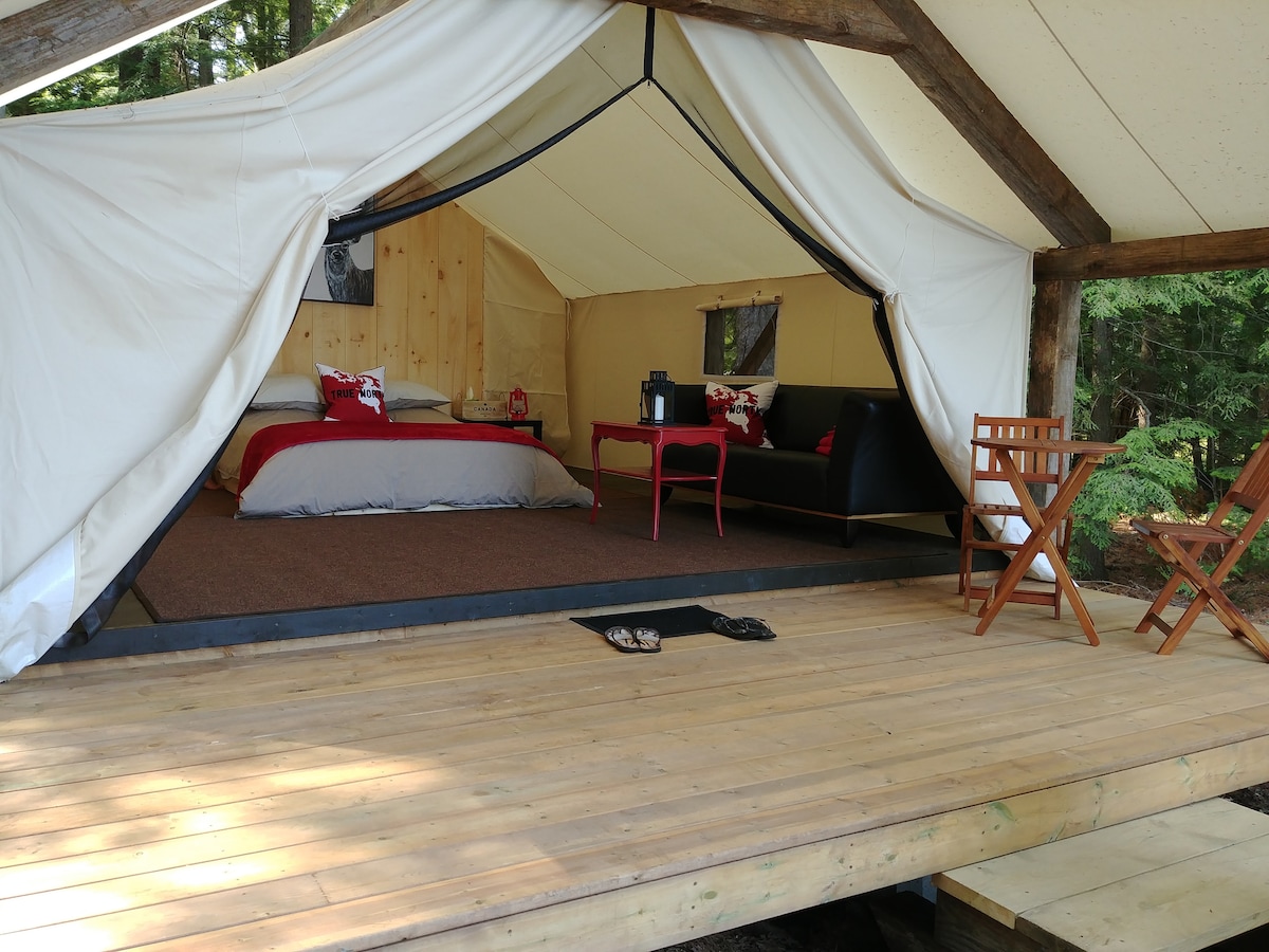 The image captures the interior of a spacious glamping tent with a raised wooden deck. A queen bed is adorned with cozy linens, accompanied by a seating area featuring a small table and chairs. Natural light filters through the tent openings, creating a warm and inviting space.