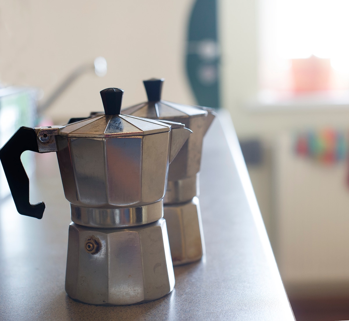 Two stovetop espresso makers are positioned on a kitchen countertop. Their metallic surfaces reflect natural light from a nearby window. A glimpse of colorful decor can be seen in the background, enhancing the kitchen's communal atmosphere.