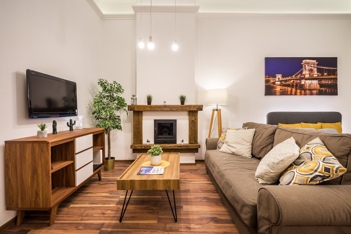 A light-filled sitting area features a comfortable sofa adorned with patterned cushions. A wooden coffee table sits centrally, complemented by a modern television mounted on the wall. A decorative fireplace and a leafy plant add warmth to the space, enhanced by soft lighting.