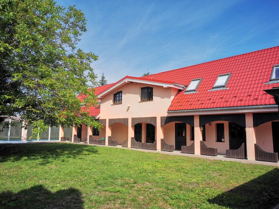A large, peach-colored building with a red tiled roof is set against a clear blue sky. The structure features arched doorways and a spacious lawn, with trees providing natural shade in the foreground.