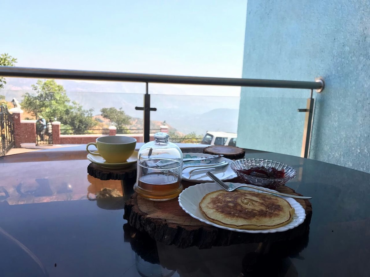 A wooden table set outdoors displays a breakfast arrangement. A plate with a pancake, a small bowl of jam, and a glass of tea is visible alongside a cup on a saucer. The background showcases a scenic view of mountains and trees, creating a serene dining atmosphere.