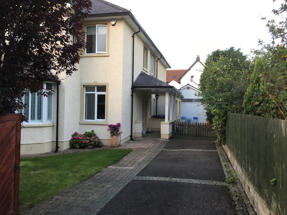 A charming exterior view of a two-story house is presented, featuring a well-maintained garden and driveway. Flower pots add a touch of color by the entrance, while a wooden gate leads to a pathway lined with decorative stones. Lush greenery surrounds the area.