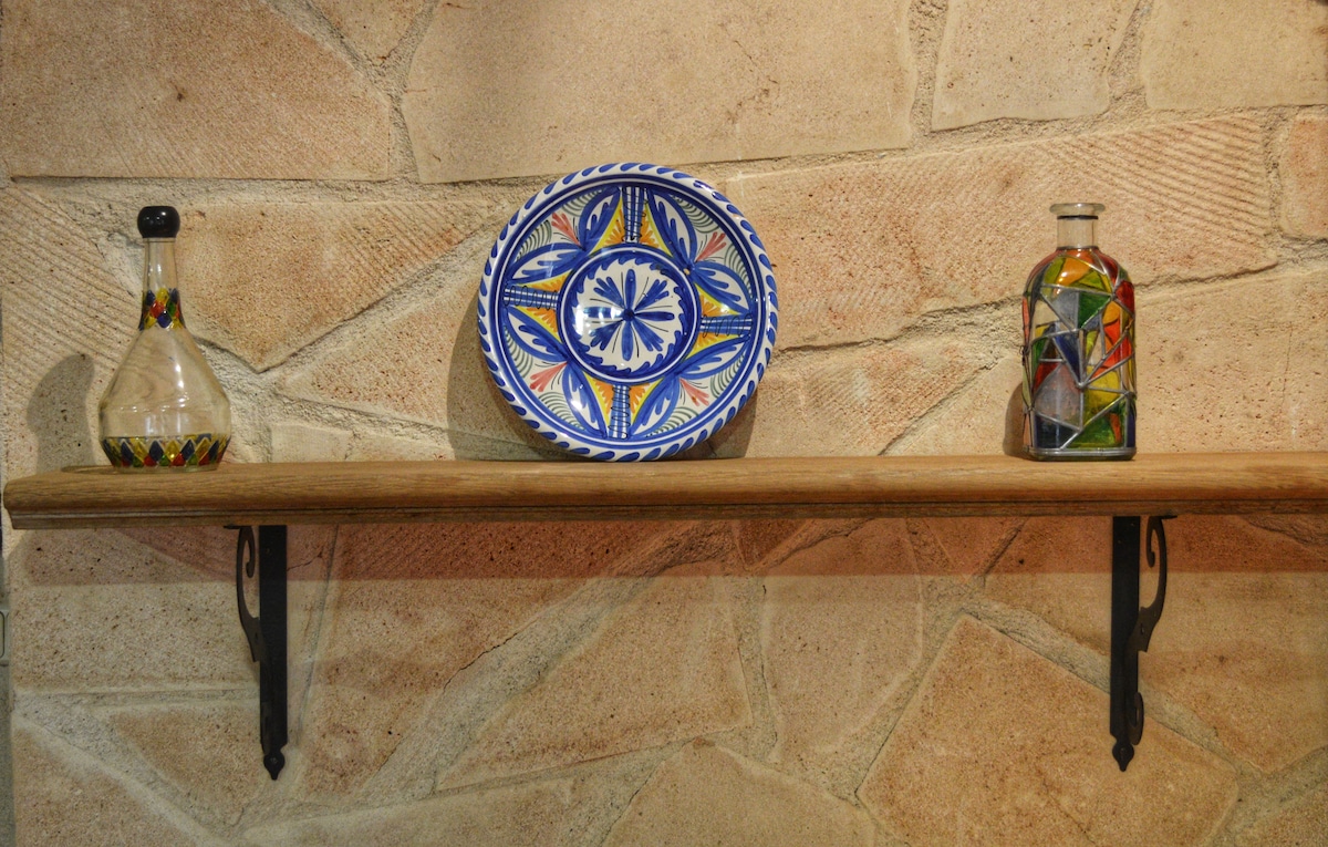 A wooden shelf rests against a textured stone wall, showcasing a decorative plate with a colorful pattern, alongside two unique glass bottles. The items feature distinct designs contributing to the overall rustic charm of the space.
