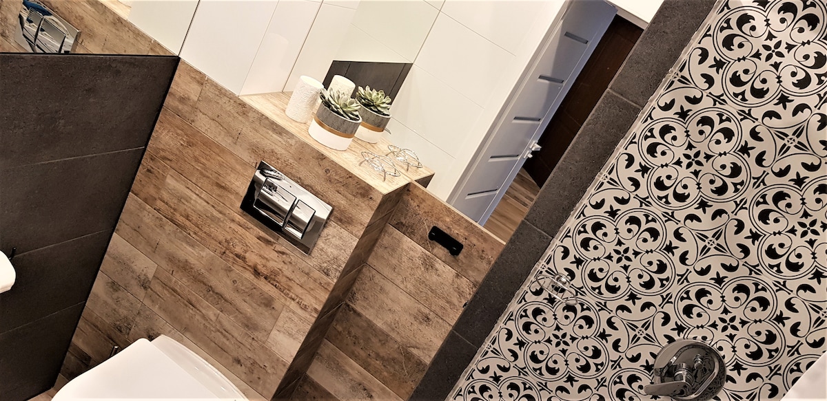 The bathroom features a combination of modern and rustic elements, with a wooden accent wall and patterned tiles. A sleek toilet is positioned next to the decorative wall, while a small countertop displays plants and glassware. Ample natural light reflects off the surfaces.