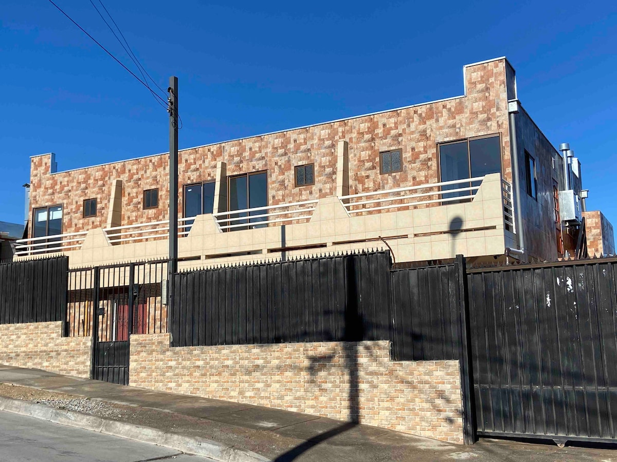 A modern two-story building is displayed, featuring a blend of textured stone and smooth materials. Large windows provide natural light to the interior. A gated entrance and fence surround the property, contributing to a sense of privacy and security.