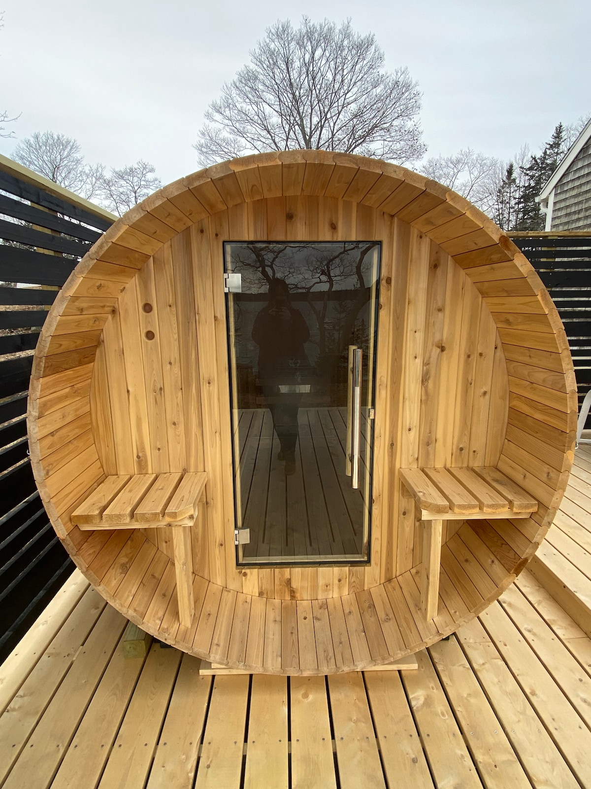 A wooden sauna featuring a circular design with a glass door, positioned on a spacious deck. Two wooden benches are placed inside, and the surrounding area includes a mix of natural wood and black privacy panels. Tall trees can be seen in the background.