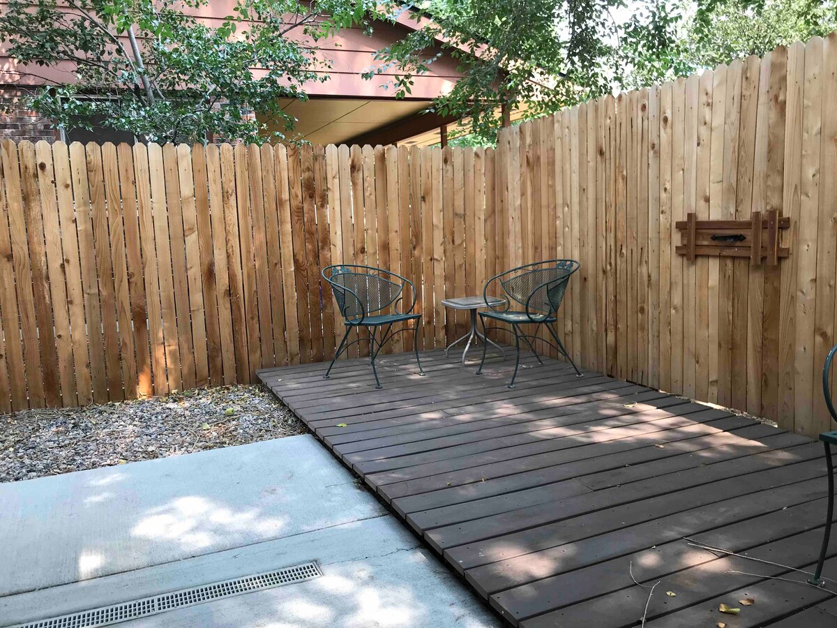 A private patio features a wooden deck surrounded by a wooden fence, offering a secluded outdoor space. Two wire chairs are positioned around a small table, providing a cozy area for relaxation. Natural elements add warmth to this tranquil setting.