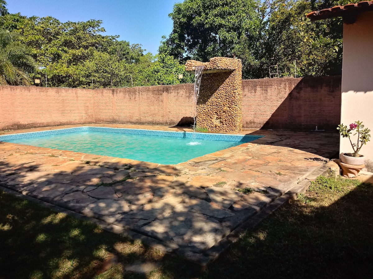 A refreshing swimming pool is set amidst tranquil greenery, featuring a natural stone waterfall that adds a serene ambiance. The surrounding area is paved with textured stones, and the softly lit edges create a peaceful outdoor space for relaxation.