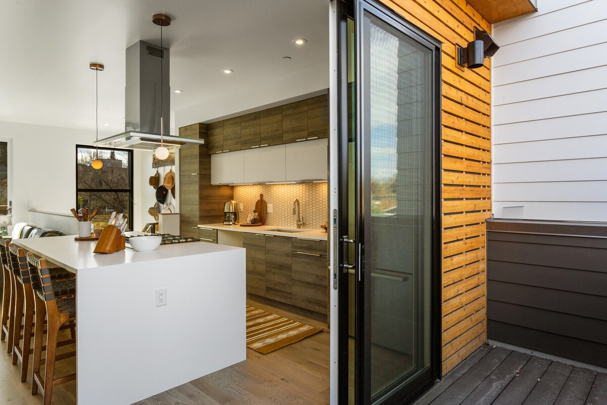 A modern kitchen features sleek cabinetry and stainless steel appliances, with an island equipped for cooking and socializing. Large glass doors open onto a deck, allowing natural light to flow through and offering a seamless connection to outdoor space.