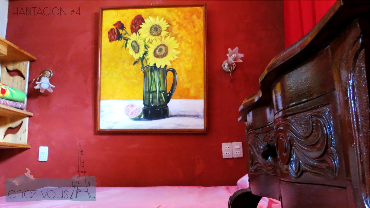 A vibrant painting of sunflowers and roses is displayed against a warm orange wall. A wooden dresser with intricate carvings is positioned nearby, complementing the inviting tones of the room. Soft lighting fixtures illuminate the space, creating a cozy ambiance.