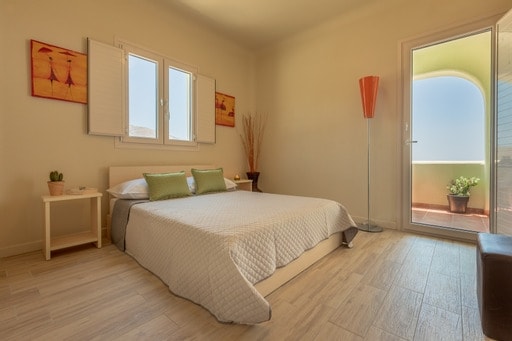 A serene bedroom is furnished with a spacious bed adorned with decorative pillows. Light wood flooring complements the warm beige walls. A bright lamp stands beside the bed, and a small table holds a potted plant. Large windows offer natural light and views of the outdoors.