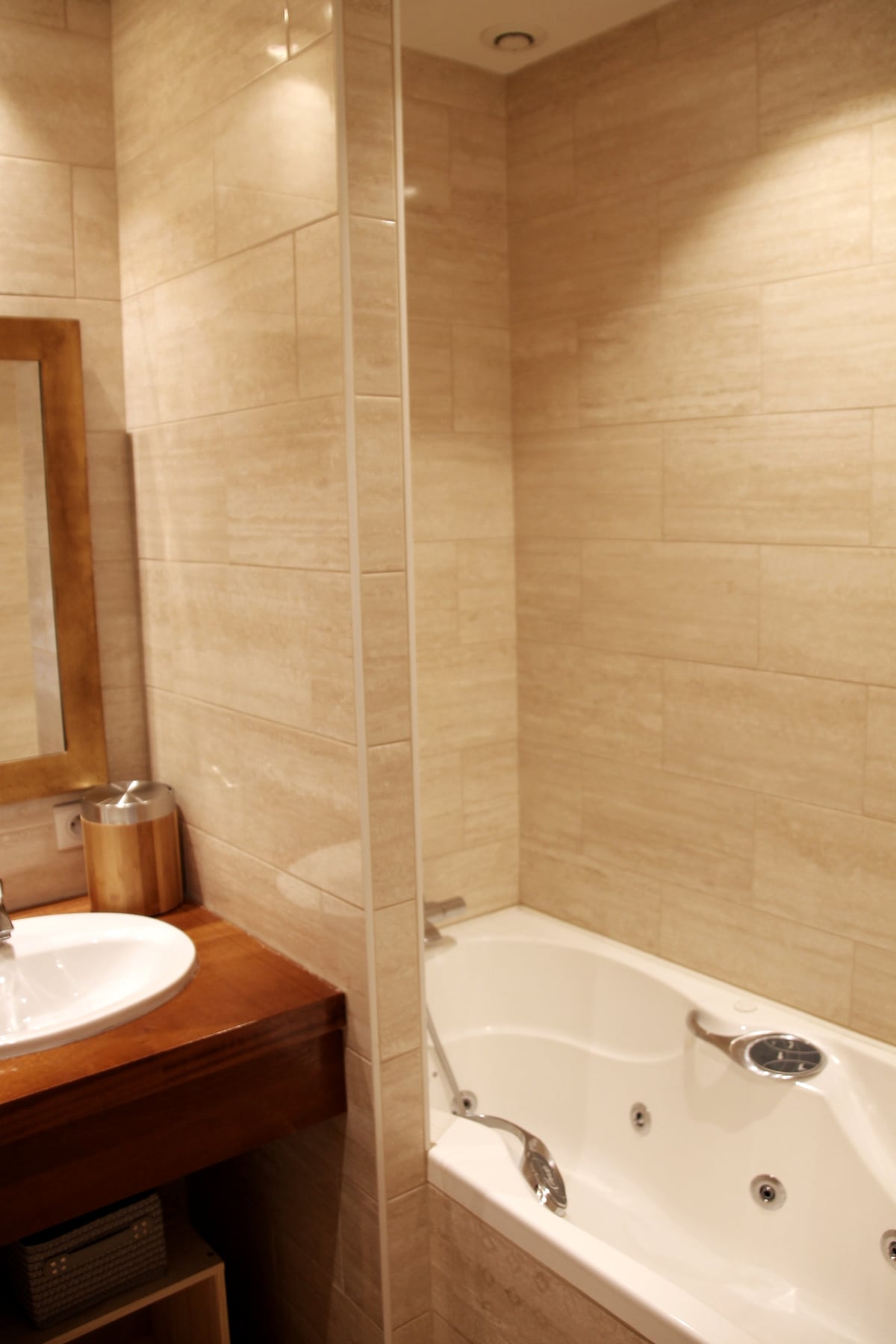 A well-appointed bathroom features a modern bathtub with a shower attachment and a glass partition. A circular sink sits atop a wooden cabinet, accompanied by a large mirror framed in wood. Soft lighting highlights the neutral tones of the tiled walls.