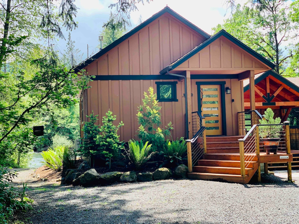 The cabin's exterior showcases a modern design with warm wood accents and a welcoming entryway. Lush greenery surrounds the structure, including ferns and small trees. A set of stairs leads to the main entrance, enhancing accessibility to this tranquil riverfront location.